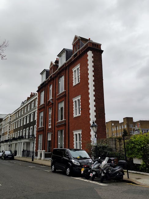 A tall, red-brick residential building with white window frames and decorative white accents running vertically along the corner, situated on a street in South Kensington. The building has multiple floors, with dormer windows in the attic space. In front of the building, a black van and several scooters are parked on the pavement, and a lamppost is visible nearby. The street scene is under an overcast sky, and other similar multi-storey buildings with Victorian architectural features line the street, reflecting typical urban housing. The image depicts a quiet residential area suitable for home relocation and furniture transport services, as provided by [COMPANY_NAME] offerring removals, with clear emphasis on urban moving logistics.