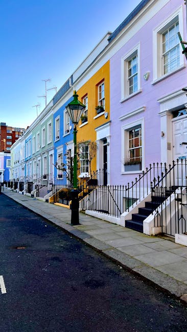 A row of pastel-colored terraced houses in South Kensington, London, featuring white-framed windows, black wrought iron fences, and small front steps. The street is paved with asphalt, with a classic black lamppost situated on the pavement. The houses display a variety of bold colors including yellow, lavender, pale blue, and light green, and are illuminated by natural daylight under a clear blue sky. This setting exemplifies typical urban residential architecture associated with house removals and home relocation services, reflecting the importance of careful planning during furniture transport and moving logistics. The image captures the quiet, well-maintained street environment where professional removal companies like [COMPANY_NAME] would organize the loading process, including loading furniture, boxes, and appliances onto removal vans, with attention to efficient and secure packing during a home move.