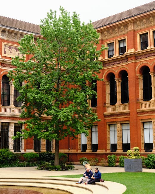 Two individuals sit on the edge of a circular stone feature outside a large red-brick building with classical architectural details, including arched windows and decorative columns. They are surrounde