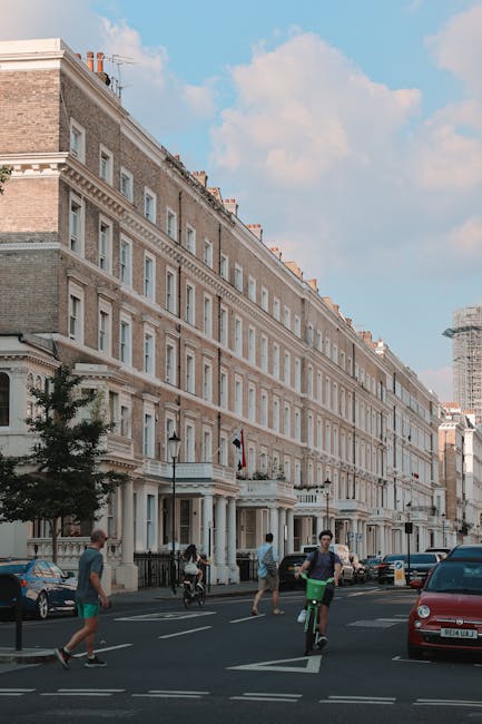 A wide street scene in a London neighbourhood with a large, multi-storey residential building featuring classic architectural details, including decorative window frames and cornices, made of light-colored brick and stone. Several pedestrians are walking on the pavement, and two individuals are riding bicycles along the road. Parked cars are lined up on the side of the street, and the sky above is partly cloudy with patches of blue visible. In the foreground, part of a moving van with open rear doors is visible, suggesting a house relocation process. The van is parked close to the building, and packaging materials such as cardboard boxes and furniture coverings are seen inside. This image reflects a typical scene during professional removals and furniture transport, with the presence of moving equipment and active loading or unloading inside the property managed by a removal company specializing in house removals near South Kensington.