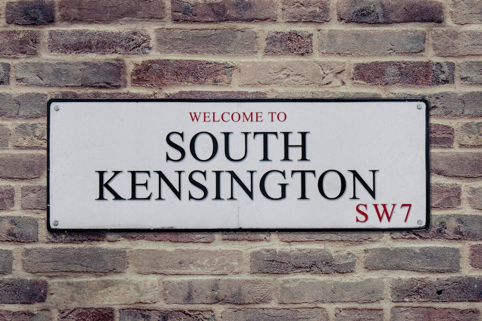A rectangular metal street sign mounted on a brick wall, displaying the words 'Welcome to South Kensington' in black uppercase letters, with the postcode 'SW7' in red at the bottom right corner. The sign is positioned indoors or on an exterior brick wall, which has a mixture of reddish-brown and beige bricks arranged in a traditional running bond pattern. The photograph highlights the sign as part of a move or relocation theme, emphasizing the coastal or city district of South Kensington, relevant to house removals or furniture transport services provided by a professional removals company.