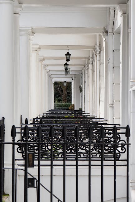 A wide street scene in a London neighbourhood with a large, multi-storey residential building featuring classic architectural details, including decorative window frames and cornices, made of light-colored brick and stone. Several pedestrians are walking on the pavement, and two individuals are riding bicycles along the road. Parked cars are lined up on the side of the street, and the sky above is partly cloudy with patches of blue visible. In the foreground, part of a moving van with open rear doors is visible, suggesting a house relocation process. The van is parked close to the building, and packaging materials such as cardboard boxes and furniture coverings are seen inside. This image reflects a typical scene during professional removals and furniture transport, with the presence of moving equipment and active loading or unloading inside the property managed by a removal company specializing in house removals near South Kensington.
