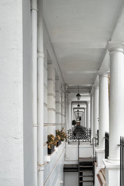 A narrow, white-painted street-facing apartment corridor with a row of tall, rounded classical columns supporting a ceiling. Several black wrought-iron outdoor light fixtures hang from the ceiling along the corridor. Along the left side, small potted plants with dried or fading foliage are placed on a ledge beneath the windows, which are framed by white masonry walls. The right side features a black metal gate or barrier at the staircase entrance, with visible steps leading downward. The space appears to be part of a residential building in South Kensington, with the corridor providing access to the street below. The environment is well-lit by natural light, emphasizing the clean, tidy appearance of the area, which could be part of a home undergoing a move or furniture transport involving multi-story access challenges, as referenced by the 'Narrow street challenges' context.