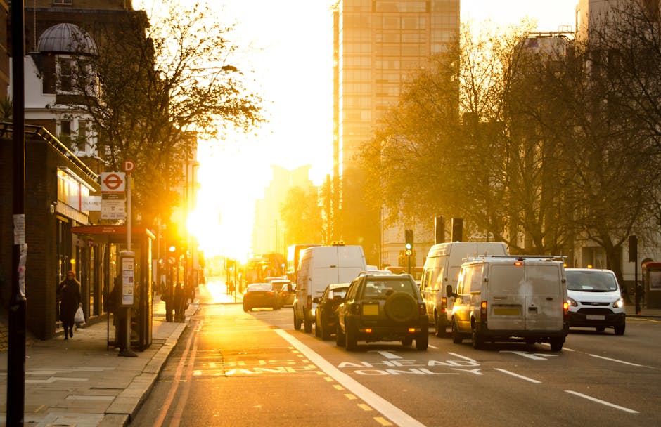 A busy urban street scene at sunset during a house removal in South Kensington, with several white and dark-colored vans and cars parked along the roadside, some being used for furniture transport and loading. In the foreground, a partially visible blue dolly and moving blankets are placed on the pavement near a group of cardboard boxes, plastic wrapping, and packing materials indicating packing and moving preparations. Pedestrians are walking on the sidewalk, and the street is lined with leafless trees and traditional London-style buildings, including a small bus shelter with a person waiting nearby. The bright sunlight creates a warm glow, casting long shadows across the scene, typical of an emergency same-day move managed by [COMPANY_NAME], involving the loading process, transportation, and logistical arrangements for home relocation within South Kensington. The environment suggests a professional move with a focus on efficient loading, packing, and furniture transport during an active moving day.