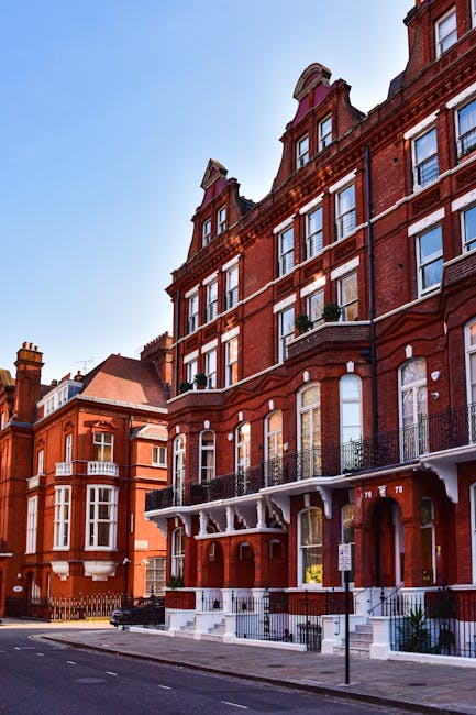 Photograph of a row of elegant Victorian-style terraced townhouses in South Kensington, London, taken during daytime with clear blue skies and scattered clouds. The buildings have light-colored brick facades with decorative cornices and tall sash windows. In the foreground, the street features parked cars, including a red hatchback and a silver sedan, with a few pedestrians and a cyclist riding along the pavement. Some of the houses have small front gardens with black iron railings and lampposts lining the sidewalk. The scene captures everyday city life in South Kensington, illustrating the setting where [COMPANY_NAME] might assist with house removals, packing, and furniture transport, emphasizing the area’s characteristic architecture and busy street environment.