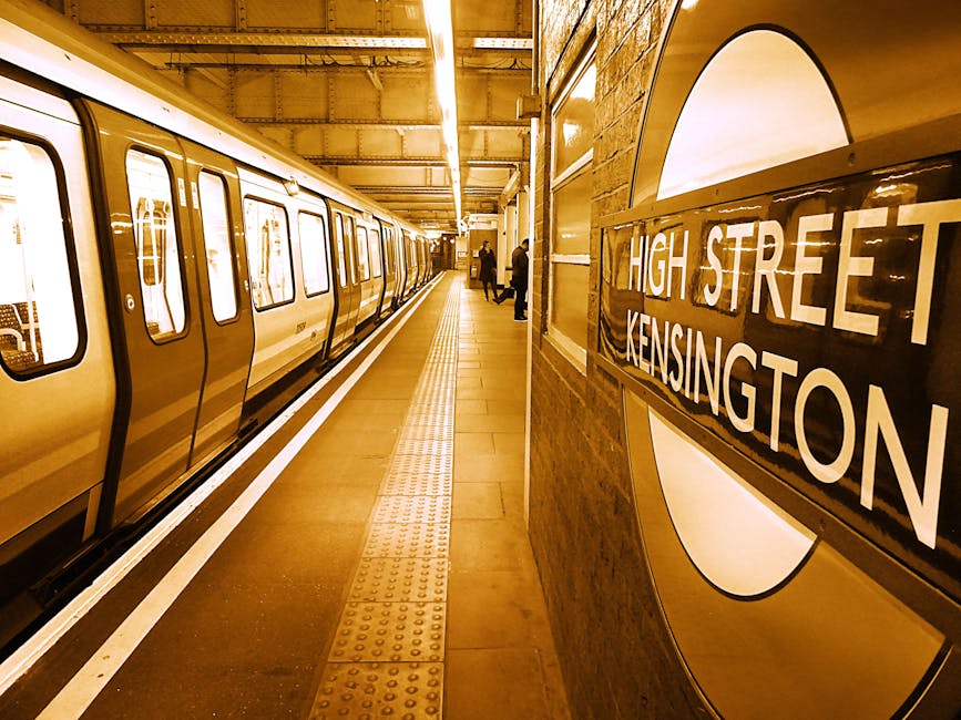 Inside the London Underground station at South Kensington, the platform is visible with a yellow train parked alongside. The train has large rectangular windows and sliding doors in the closed position. The platform surface is made of light brown tiles with tactile paving strips near the edge for safety. Several people are standing on the platform, some facing towards the tunnel, others walking or waiting. On the right side of the image, a wall features a large round London Underground sign with the station name 'High Street Kensington' displayed in white letters on a black background. The station's lighting is warm, casting a yellowish glow over the scene. This setting illustrates the typical environment of a busy urban underground station, relevant for home relocation or moving logistics when planning transportation options in South Kensington.