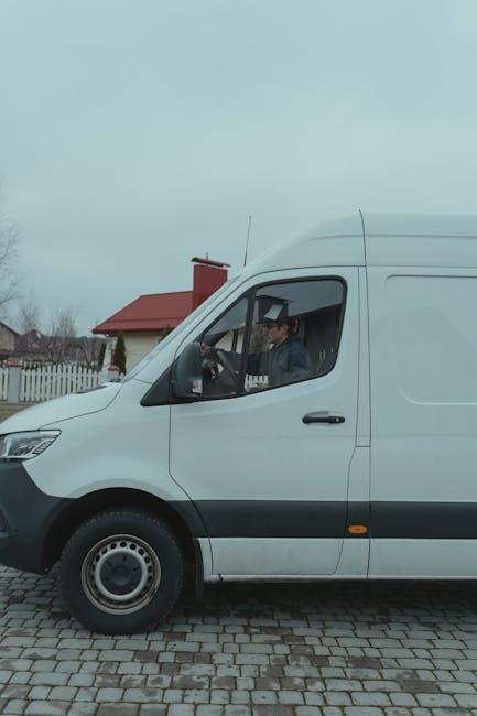 A white commercial van of the type used in house removals and furniture transport is parked on a cobblestone driveway. The van has a high roof and sliding side door, with a driver visible through the window, wearing a cap and dark clothing. The surrounding environment includes a residential area with a house featuring a red roof and a chimney, a white picket fence, and bare trees, indicating an outdoor setting in a suburban neighbourhood. The van is positioned near a doorway, suggesting it is engaged in loading or unloading furniture and boxes for a house move. The image captures the loading process typical of professional removals, highlighting the importance of careful packing and transportation during home relocations, as managed by companies like [COMPANY_NAME]. The lighting appears natural, with overcast skies, and the scene emphasizes organized, efficient moving logistics involving vans, packaging materials, and mobility equipment like trolleys or straps.