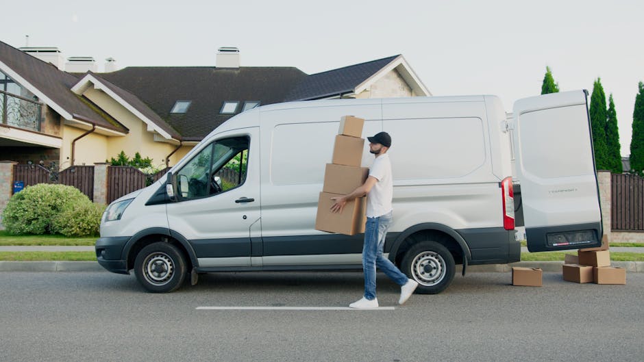 A white commercial van of the type used in house removals and furniture transport is parked on a cobblestone driveway. The van has a high roof and sliding side door, with a driver visible through the window, wearing a cap and dark clothing. The surrounding environment includes a residential area with a house featuring a red roof and a chimney, a white picket fence, and bare trees, indicating an outdoor setting in a suburban neighbourhood. The van is positioned near a doorway, suggesting it is engaged in loading or unloading furniture and boxes for a house move. The image captures the loading process typical of professional removals, highlighting the importance of careful packing and transportation during home relocations, as managed by companies like [COMPANY_NAME]. The lighting appears natural, with overcast skies, and the scene emphasizes organized, efficient moving logistics involving vans, packaging materials, and mobility equipment like trolleys or straps.